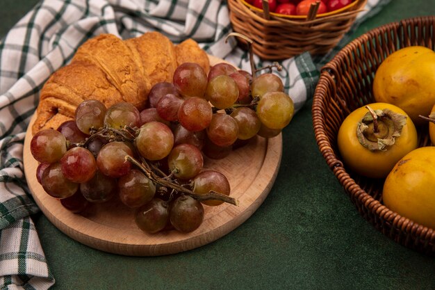 Vista superior de las uvas en una tabla de cocina de madera sobre un paño de cuadros con croissant con frutas de caqui en un balde sobre un fondo verde