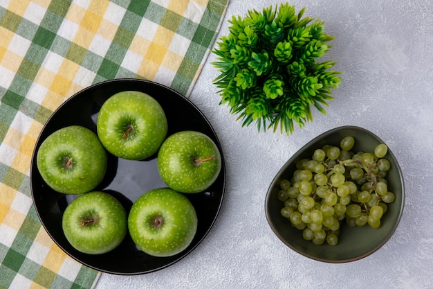 Vista superior de uvas verdes en un tazón de manzanas verdes con una toalla a cuadros verde-amarillo sobre un fondo blanco.