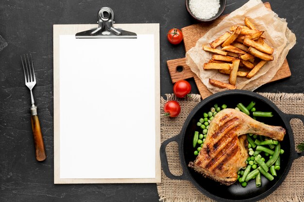 Vista superior de vainas de pollo y guisantes al horno en sartén con patatas y portapapeles en blanco