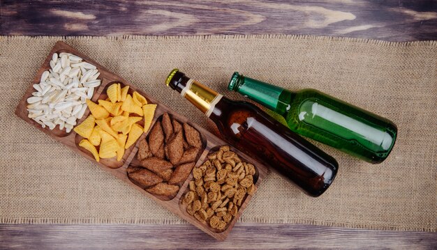 Vista superior de variados aperitivos de cerveza, galletas saladas, papas fritas y semillas de girasol en una bandeja de madera con botellas de cerveza en tela de saco rústico