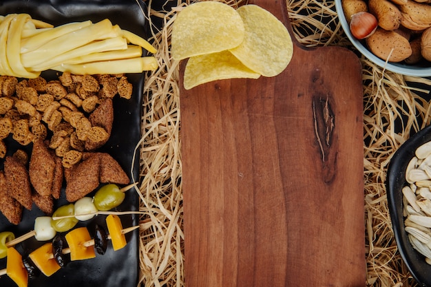 Vista superior de variados aperitivos de cerveza papas fritas aceitunas en vinagre galletas saladas de pan y queso y una tabla para cortar madera en paja