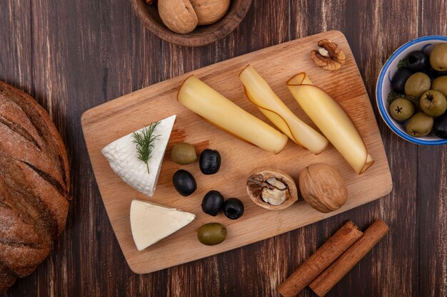 Vista superior de variedades de queso y aceitunas en un soporte con canela y hogazas de pan sobre un fondo de madera