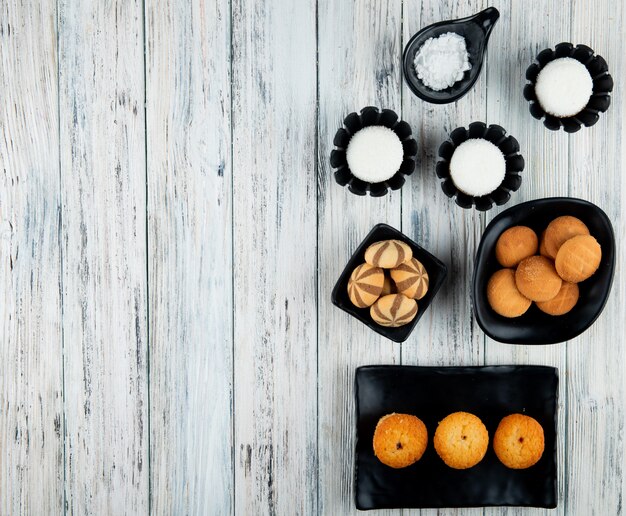 Vista superior de varios tipos de galletas dulces y muffins en bandejas negras sobre fondo de madera con espacio de copia