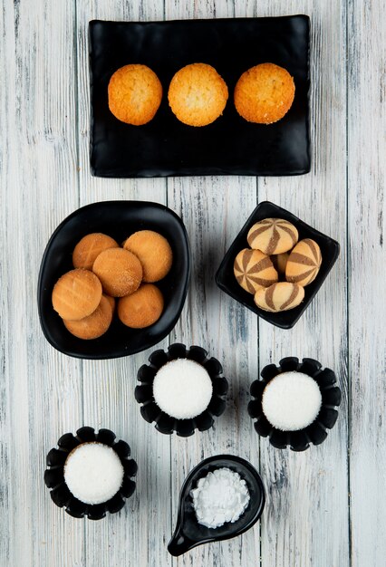 Vista superior de varios tipos de galletas dulces y muffins en bandejas negras sobre fondo de madera