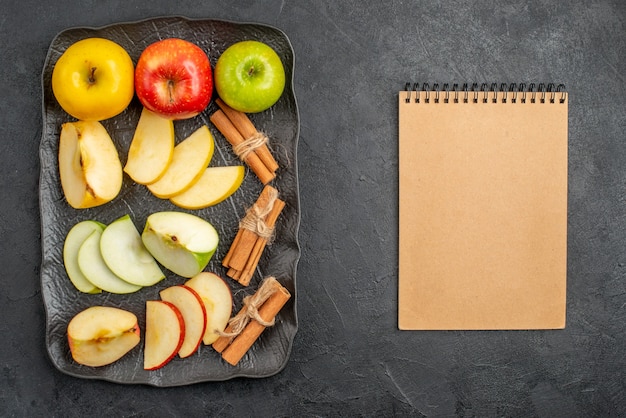 Vista superior de varios tipos de manzanas frescas en rodajas y enteras en una bandeja negra y limones canela junto al cuaderno sobre un fondo oscuro