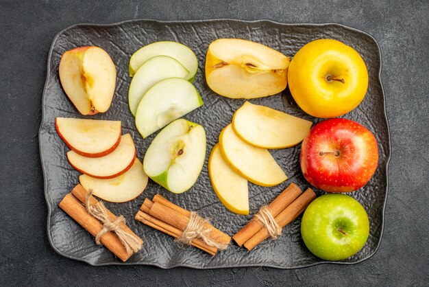 Vista superior de varios tipos de manzanas frescas en rodajas y enteras en una bandeja negra y limones canela sobre un fondo oscuro