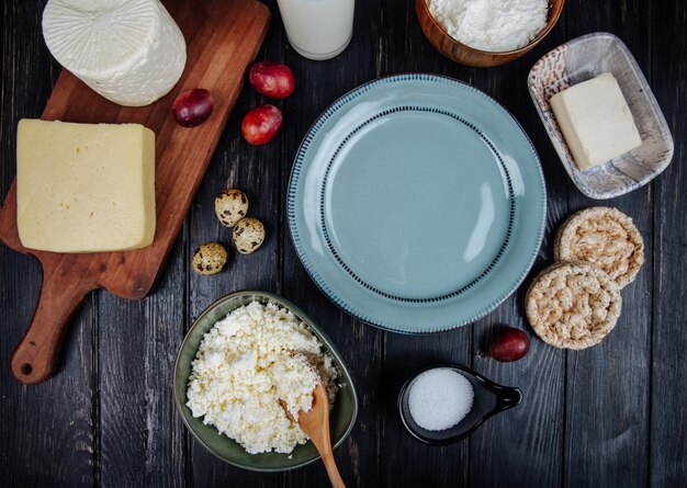 Vista superior de varios tipos de queso con requesón en un tazón, huevos de codorniz, uvas frescas dulces y un plato vacío en la mesa de madera oscura