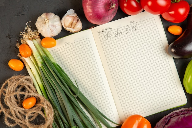 Vista superior de varios vegetales frescos tomates cebolla verde y ajo con cuaderno en el medio en la oscuridad