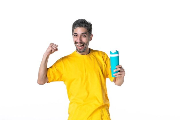 Vista superior del varón joven sonriente en camisa amarilla que sostiene el termo que se siente confiado en el fondo blanco