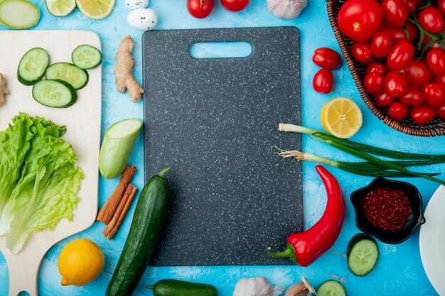 Vista superior de vegetales como lechuga, pepino, canela, jengibre y otros con corte de boarda sobre fondo azul