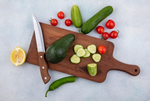 Vista superior de vegetales frescos como pepino tomate aguacate limón en tablero de cocina con cuchillo en blanco
