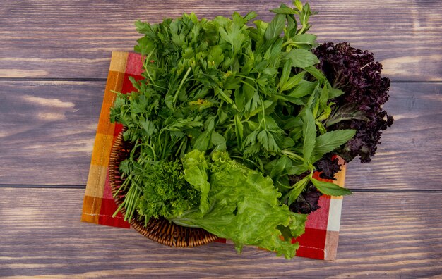 Vista superior de vegetales verdes como cilantro menta lechuga albahaca en canasta sobre tela en madera