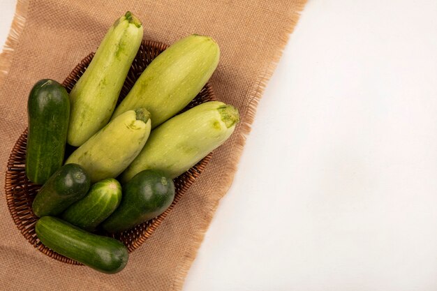 Vista superior de vegetales verdes saludables como pepinos calabacines en un cubo sobre una tela de saco sobre un fondo blanco con espacio de copia