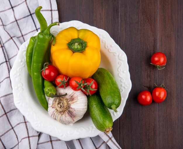 Vista superior de verduras como ajo pimiento pepino y tomate en un tazón sobre tela escocesa y superficie de madera