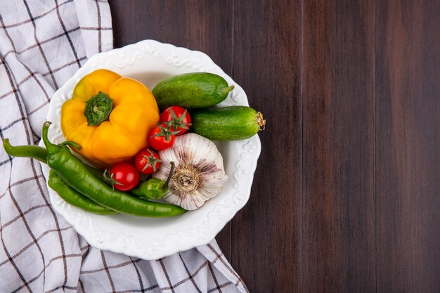 Vista superior de verduras como ajo pimiento pepino y tomate en un tazón sobre tela escocesa y superficie de madera