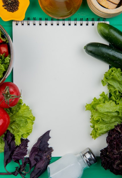 Vista superior de verduras como albahaca tomate lechuga pepino con sal y pimienta negra con bloc de notas en superficie verde con espacio de copia