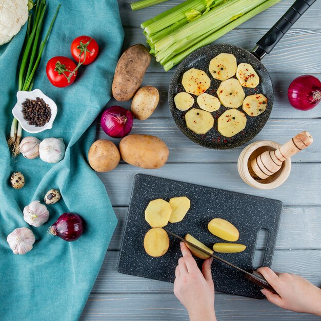 Vista superior de verduras como apio tomate cebolla ajo huevo con mujer manos cortar patatas en tabla de cortar sobre fondo de madera