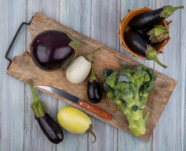 Vista superior de verduras como berenjenas y brócoli con un cuchillo en la tabla de cortar y en la canasta y sobre fondo de madera