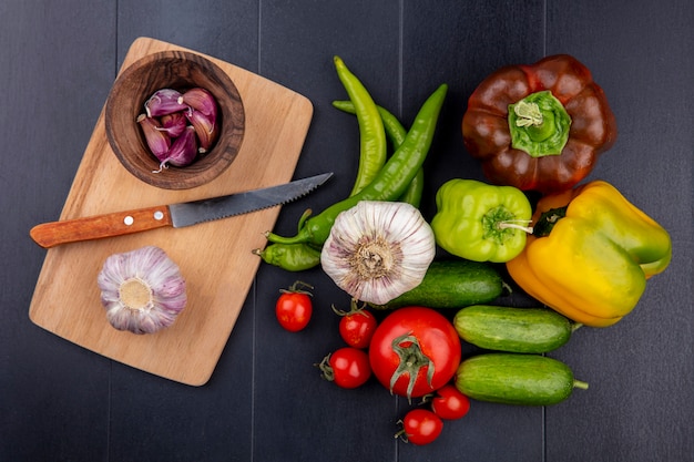 Vista superior de verduras como bulbo de ajo y dientes con cuchillo en la tabla de cortar y tomate pimiento pepino sobre superficie negra