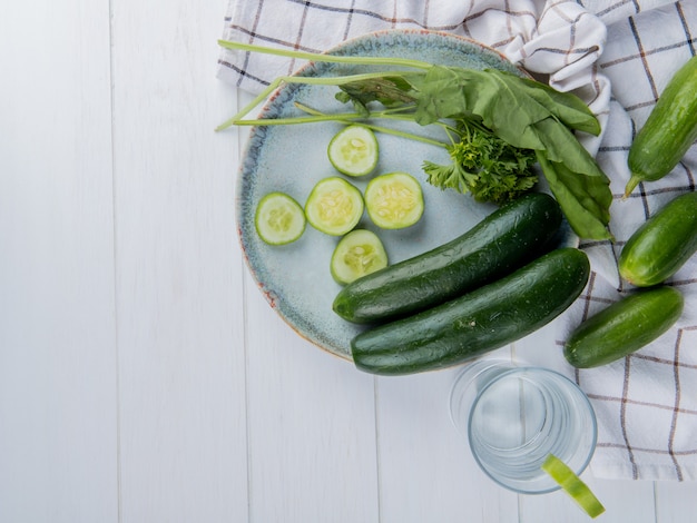 Vista superior de verduras como pepino entero y en rodajas de espinacas y cilantro con pepinos sobre tela y agua de desintoxicación en superficie de madera con espacio de copia