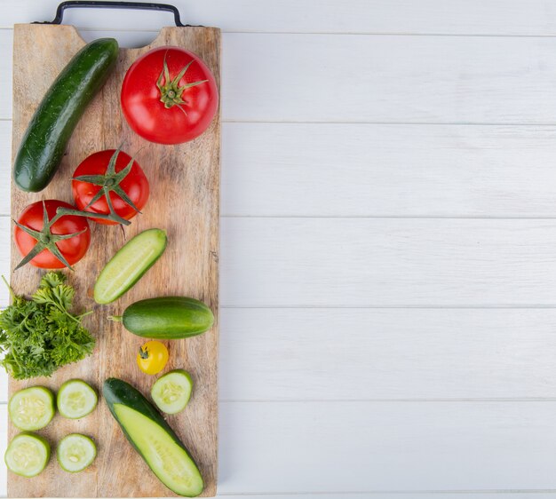 Vista superior de verduras como pepino tomate cilantro en tabla de cortar en el lado izquierdo y superficie de madera con espacio de copia