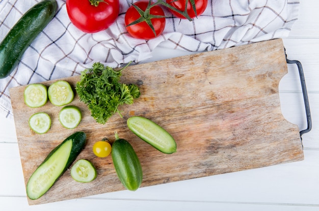 Vista superior de verduras como pepino tomate cilantro en tabla de cortar con pepino y tomate sobre tela y madera