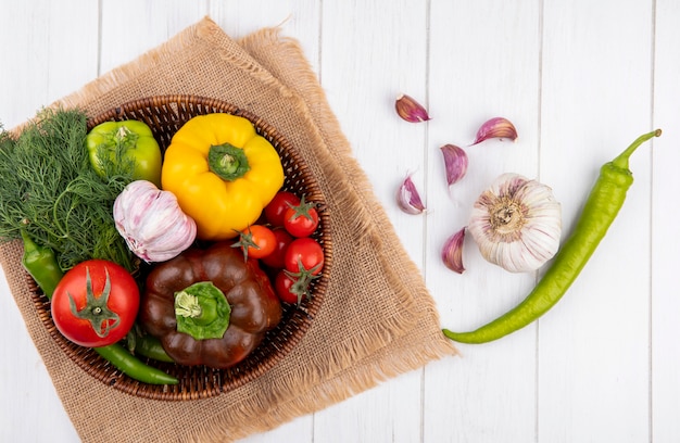 Vista superior de verduras como pimiento ajo eneldo tomate en canasta sobre tela de saco con dientes de ajo sobre superficie de madera