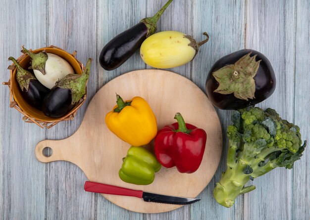 Vista superior de verduras como pimientos con cuchillo sobre tabla de cortar y berenjenas con brócoli sobre fondo de madera