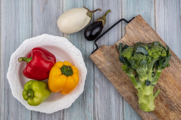 Vista superior de verduras como pimientos en un tazón y brócoli en la tabla de cortar con berenjenas sobre fondo de madera