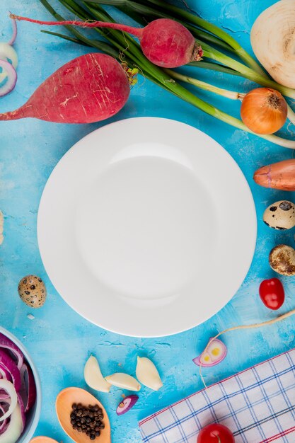 Vista superior de verduras como rábano cebolleta ajo con pimienta negra y plato en el centro sobre fondo azul.