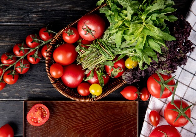 Vista superior de verduras como tomate hojas de menta verde albahaca en la cesta y corte el tomate en la bandeja sobre superficie de madera