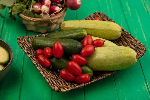 Vista superior de verduras como tomates ciruela pepinos y calabacines en una bandeja de mimbre con rábanos en un balde en una pared de madera verde