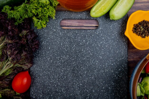 Vista superior de verduras cortadas y enteras como tomate albahaca menta pepino cilantro con pimienta negra y tabla de cortar sobre superficie de madera