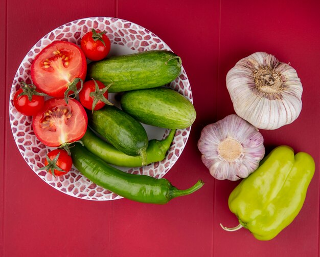 Vista superior de verduras cortadas y tomates enteros pimientos y pepinos enteros en un tazón con bulbos de ajo en superficie roja