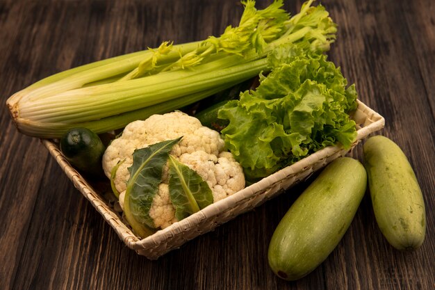 Vista superior de verduras frescas como apio lechuga y coliflor en un balde con calabacines aislado sobre un fondo de madera