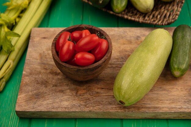 Vista superior de verduras frescas como calabacín pepino en una tabla de cocina de madera con tomates ciruela en un cuenco de madera con apio aislado en una pared de madera verde