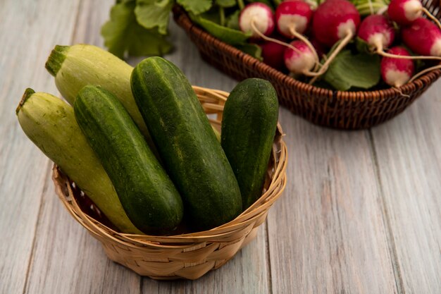 Vista superior de verduras frescas como pepinos y calabacines en un balde con rábanos en un balde sobre una pared de madera gris