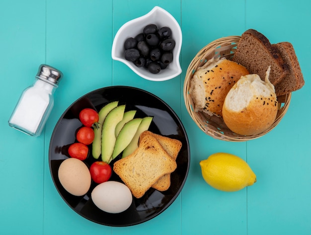 Vista superior de verduras frescas como rodajas de aguacate, tomates, aceitunas, huevos en placa negra con un cubo de panes limón en azul