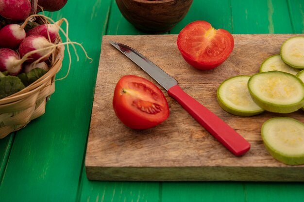 Vista superior de verduras frescas como tomates y rodajas picadas de calabacín aislado en una tabla de cocina de madera con un cuchillo con rábanos en un cubo en una pared de madera verde