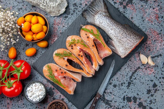 Vista superior de verduras frescas de pescado crudo en bandeja de color oscuro especias tomates con tallos ajos flores cuchillo kumquats en mesa de colores de mezcla azul negro