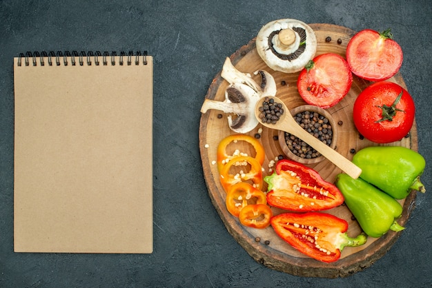 Vista superior de verduras frescas seta pimienta negra en cuchara de madera y tazón de fuente tomates rojos pimientos en el bloc de notas de tablero de madera sobre fondo oscuro