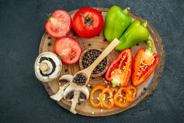 Vista superior de verduras frescas setas pimienta negra en cuchara de madera y tazón de fuente tomates rojos pimientos sobre tablero de madera sobre fondo oscuro