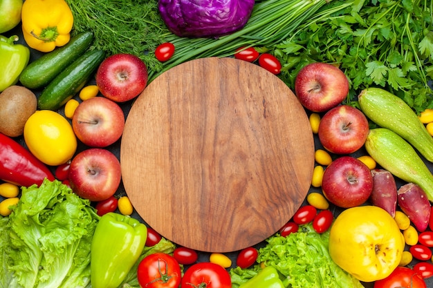 Vista superior de verduras y frutas, lechuga, tomates, pepino, eneldo, tomates cherry, calabacín, cebolla verde, perejil, manzana, limón, kiwi, tablero de madera redonda en el centro
