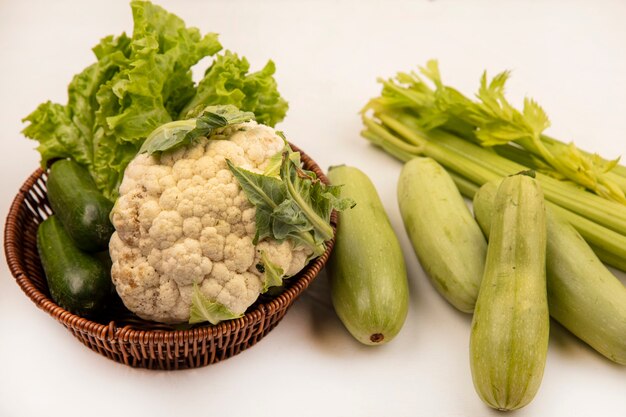 Vista superior de verduras ricas en vitaminas como lechuga, coliflor y pepinos en un balde con apio y calabacines aislados en una pared blanca