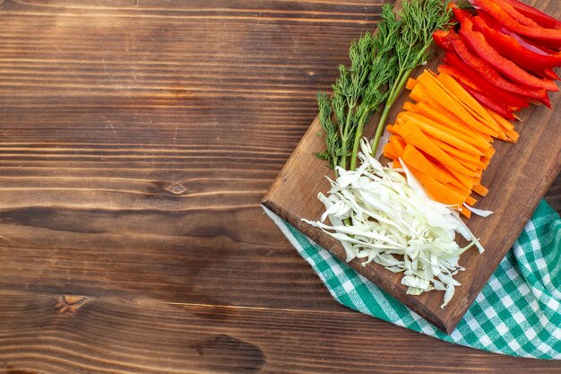 Vista superior de verduras en rodajas, repollo, zanahoria y pimiento en la superficie marrón de la tabla de cortar
