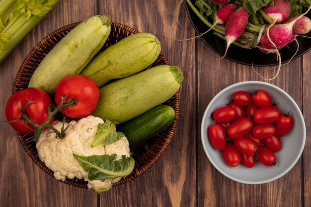 Vista superior de verduras saludables como calabacín, pepino y coliflor en un balde con tomates en un recipiente con rábanos en un plato sobre una pared de madera