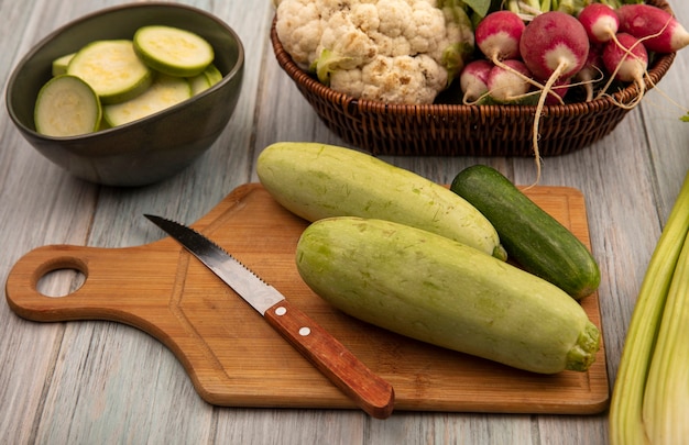 Vista superior de verduras saludables como calabacines y pepinos en una tabla de cocina de madera con un cuchillo con calabacines picados en un recipiente sobre un fondo de madera gris