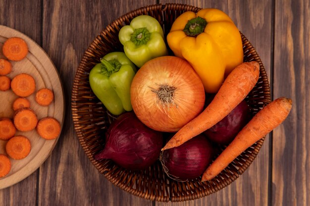 Vista superior de verduras saludables como cebollas blancas y rojas, pimientos y zanahorias de colores en un balde sobre una superficie de madera