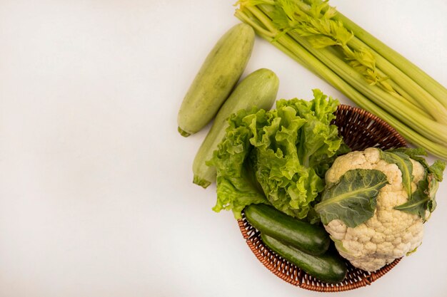 Vista superior de verduras saludables como lechuga, coliflor y pepinos en un balde con apio y calabacines aislado en una pared blanca con espacio de copia