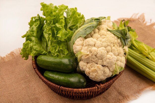 Vista superior de verduras saludables como lechuga, coliflor y pepinos en un balde sobre una tela de saco con apio aislado en una pared blanca